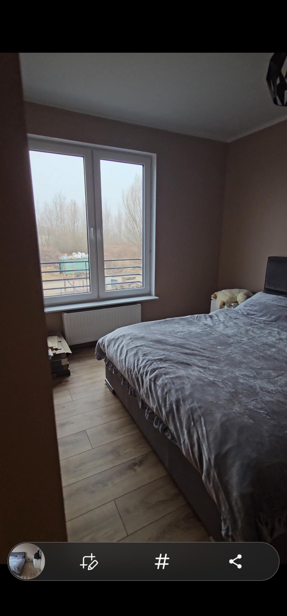 Pokój z łóżkiem i drewnopodobną podłogą, widok na zewnątrz przez okno z grzejnikiem, częściowo widoczne panele podłogowe przy ścianie.