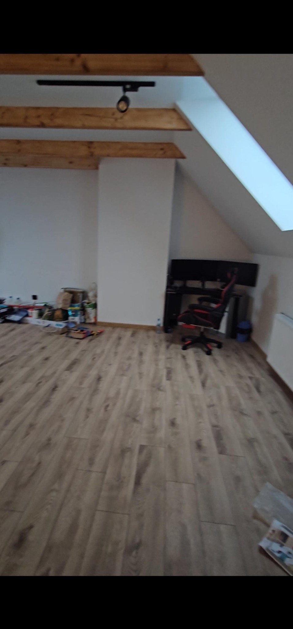 Poddasze z drewnianą podłogą panelową, widoczne drewniane belki stropowe, okno dachowe, biurko z monitorami i krzesło gamingowe, oraz pozostałości po pracach remontowych.