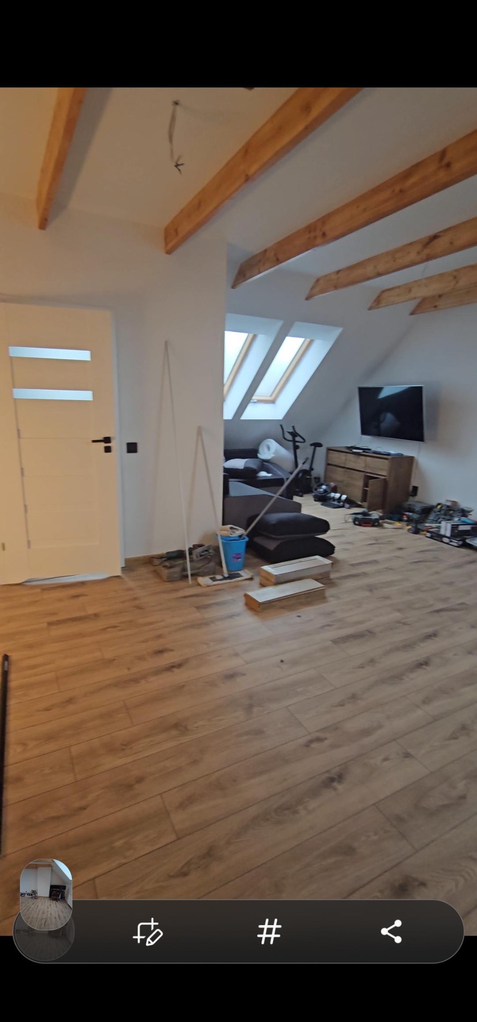 Podłoga z paneli w trakcie układania w pokoju na poddaszu, widoczne drewniane belki stropowe, białe ściany i drzwi, oraz okna dachowe. W tle widoczne sprzęty domowe i narzędzia.