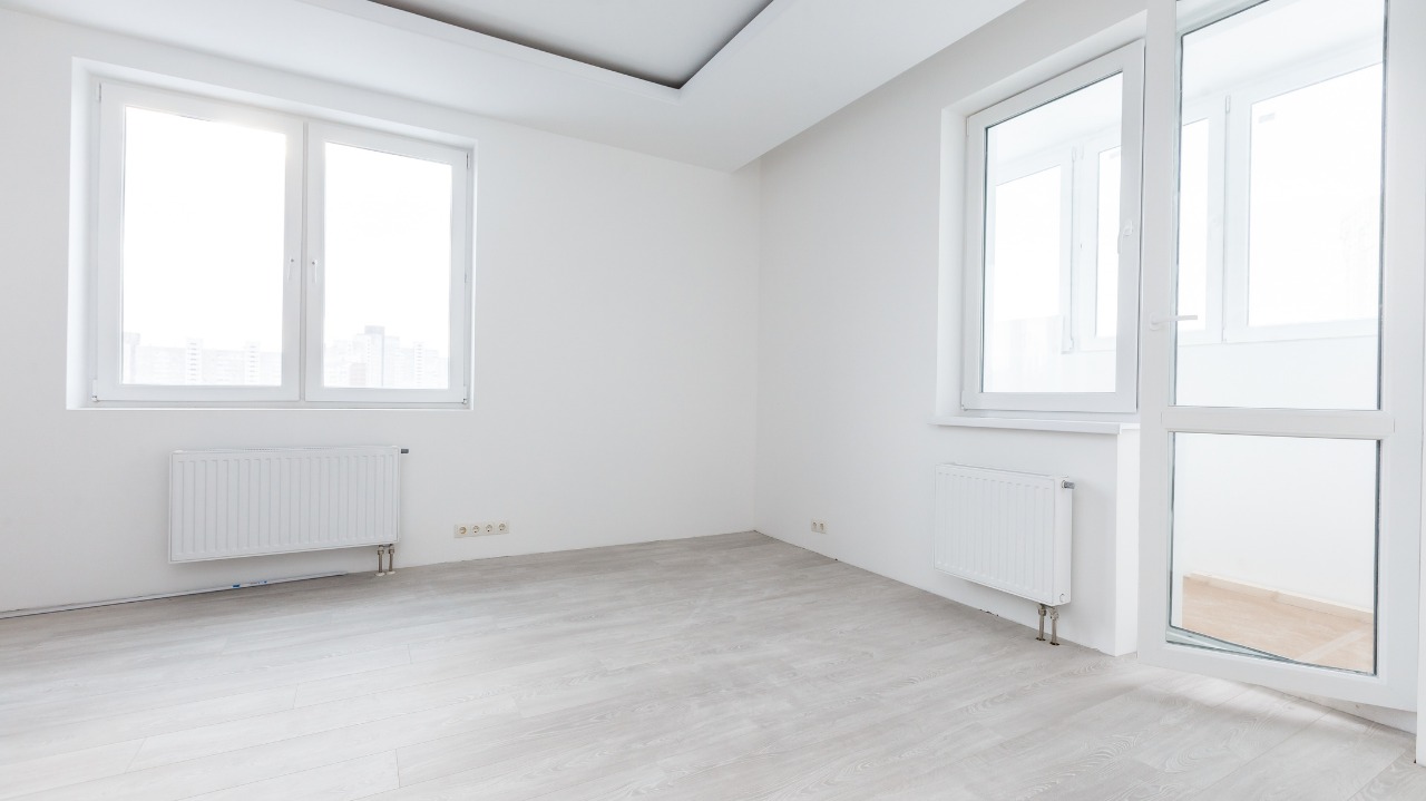 Jasne, puste pomieszczenie z panelami podłogowymi, białymi ścianami, grzejnikami pod oknami i oknem balkonowym, przygotowane do dalszego wykończenia.