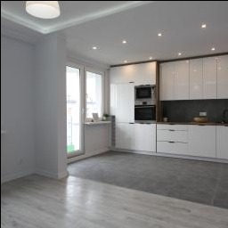 Nowoczesna, jasna kuchnia z białymi, lakierowanymi szafkami, szarym blatem i podłogą z płytek imitujących beton. Widoczne wbudowane sprzęty AGD: piekarnik i mikrofalówka. Minimalistyczne...