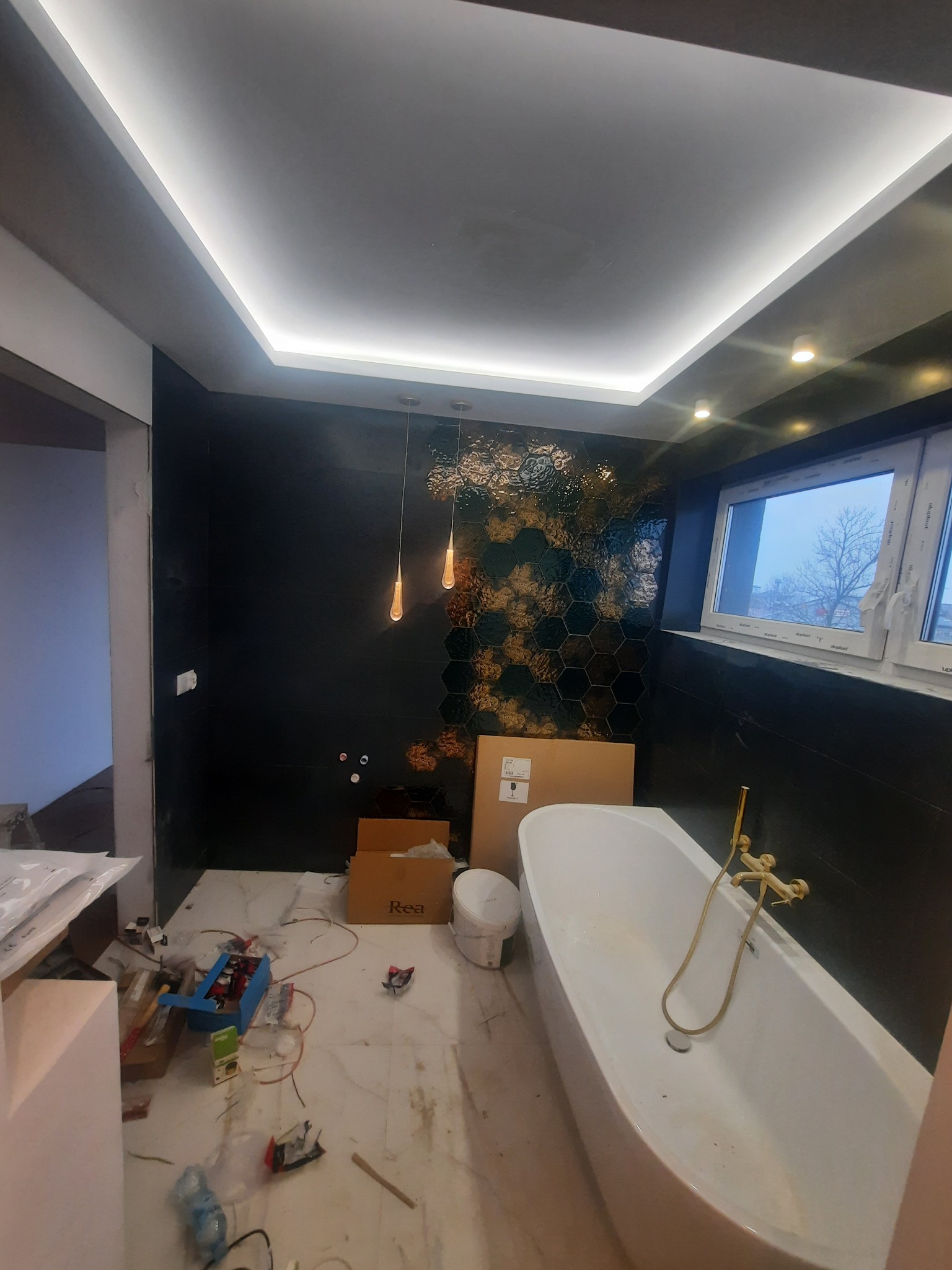 Łazienka w trakcie remontu: widoczna wanna wolnostojąca z złotą armaturą, heksagonalne płytki na ścianie, oświetlenie sufitowe LED i akcesoria budowlane na podłodze.