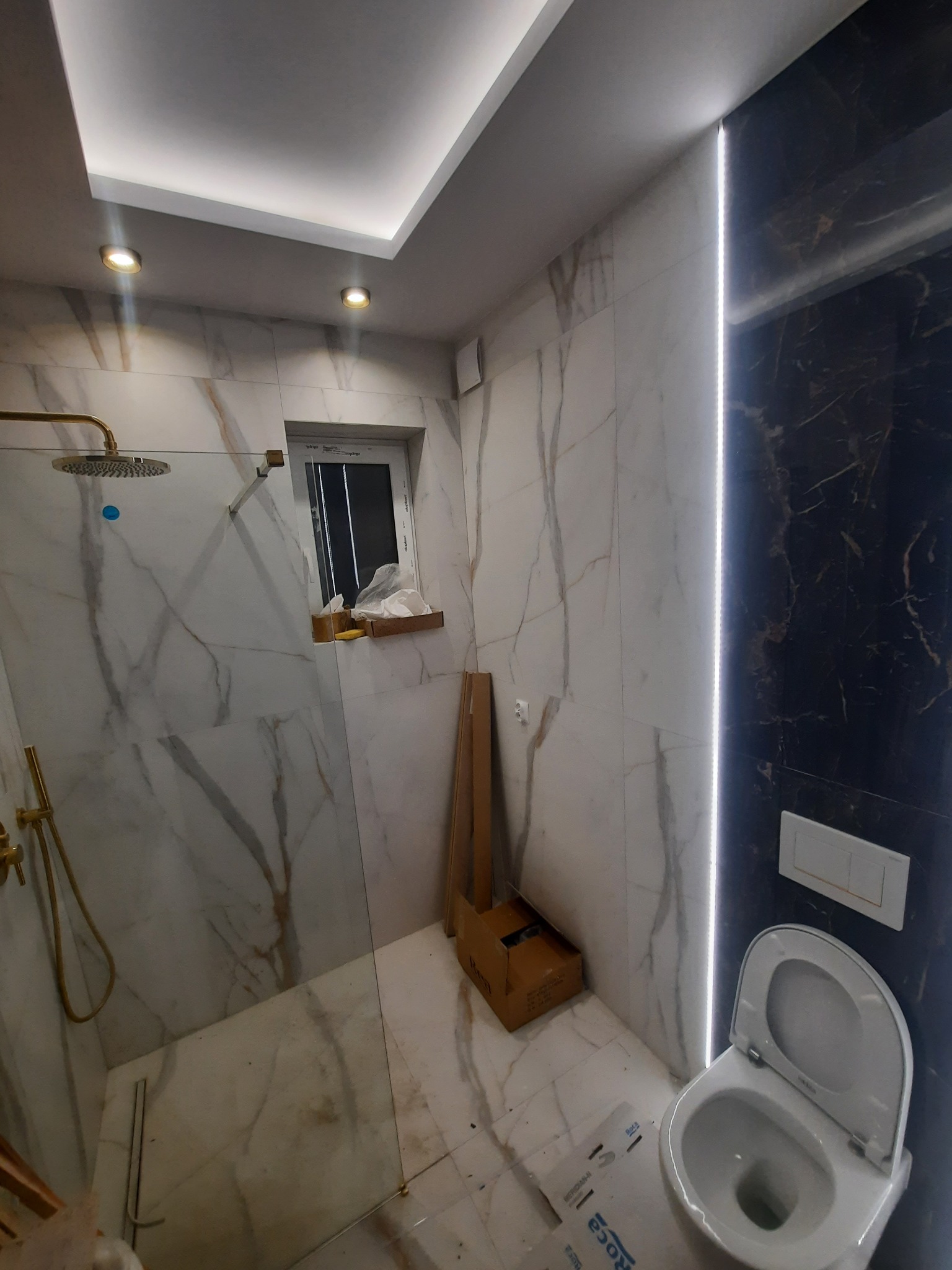 Wykończona łazienka z kabiną prysznicową typu walk-in, złotą armaturą, marmurowymi płytkami na ścianach i podświetleniem LED wzdłuż czarnej ściany, widoczna toaleta i kartony po materiałach.