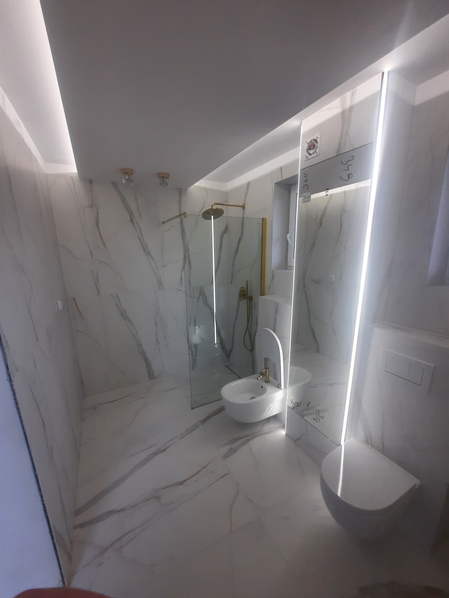 Nowoczesna łazienka wykończona białym marmurem z złotymi akcentami, kabina prysznicowa ze złotą armaturą, podświetlane lustro z widocznymi notatkami, wisząca toaleta i bidet.