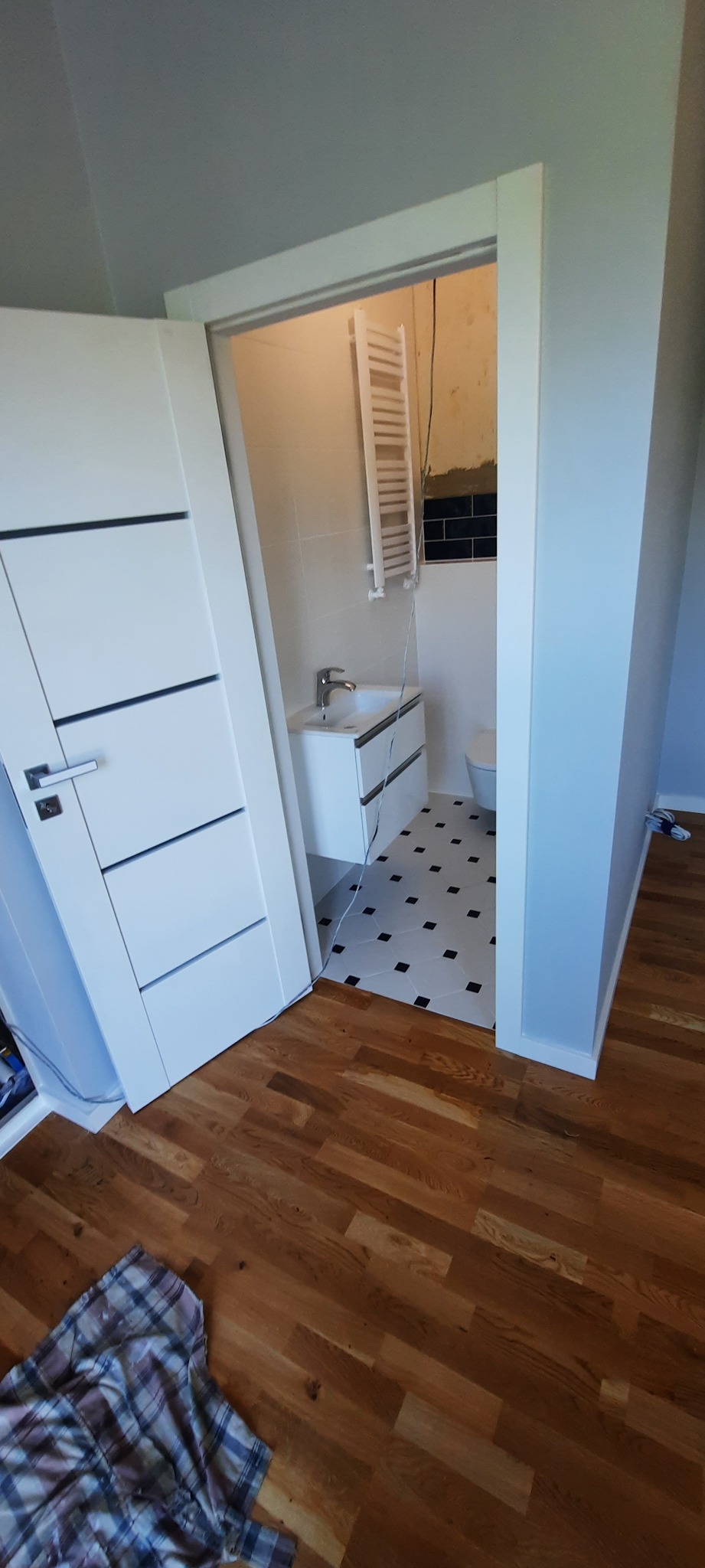Częściowo wykończona łazienka z białą umywalką na szafce, grzejnikiem drabinkowym, czarno-białą podłogą i fragmentem ściany z czarnymi płytkami, widoczna zza otwartych, białych drzwi z poziomymi...