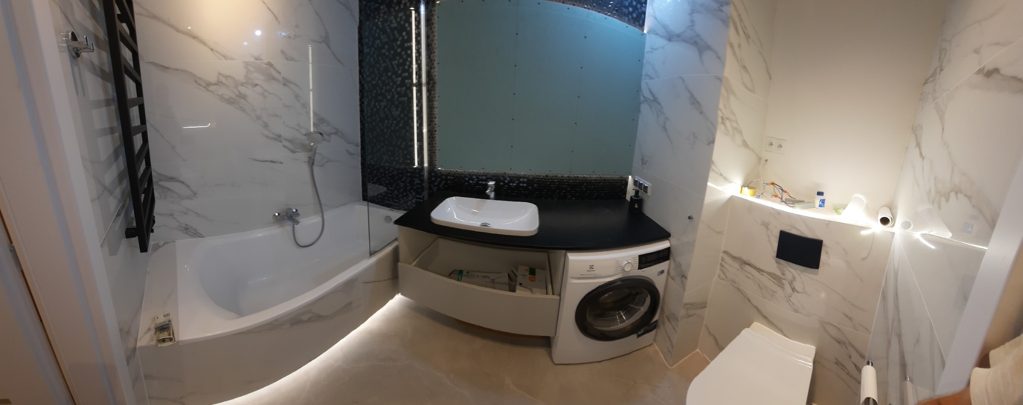 Nowoczesna łazienka z wanną obudowaną marmurową płytką, umywalką nablatową na czarnym blacie z pralką pod spodem oraz toaletą podwieszaną z podświetleniem LED.