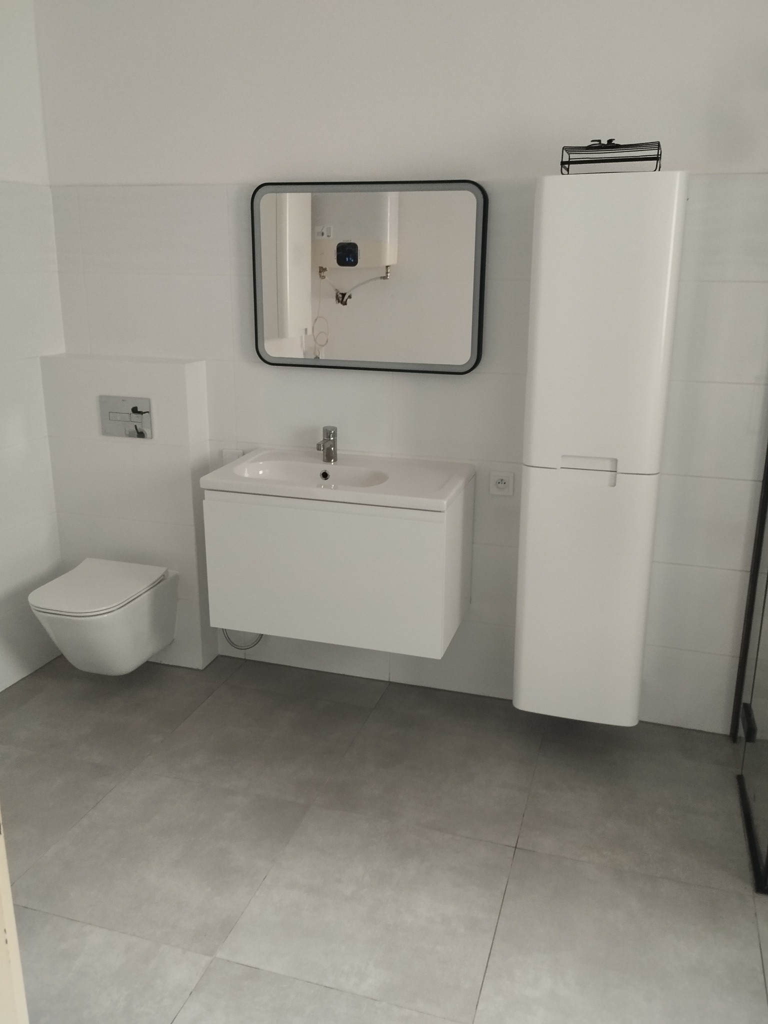 Nowoczesna łazienka w bieli i szarości: podwieszana toaleta, szafka z umywalką, lustro w czarnej ramie i wysoka szafka, szare płytki podłogowe.