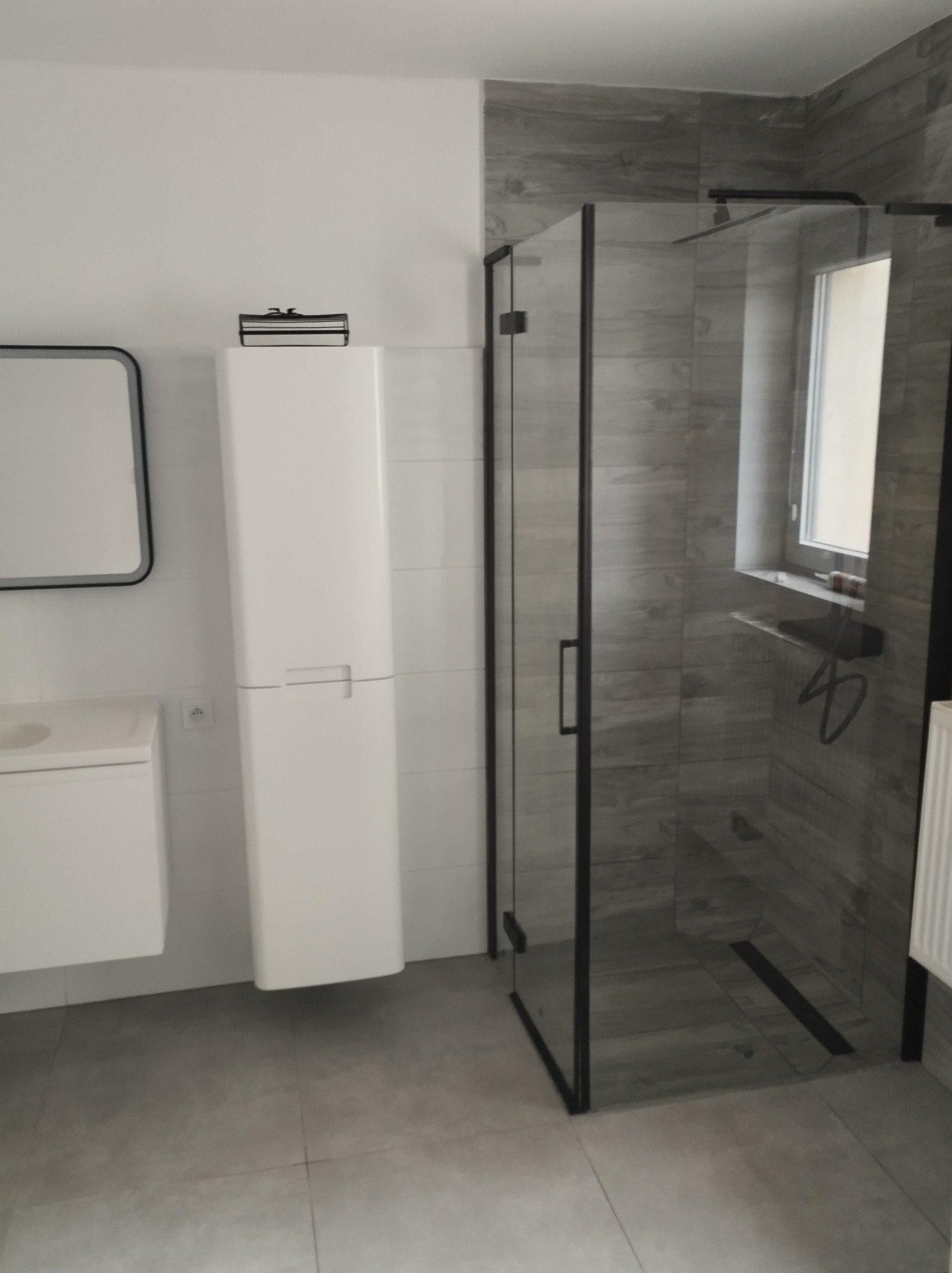 Nowoczesna łazienka z kabiną prysznicową typu walk-in z czarnymi profilami, białą szafką wiszącą i szarą płytką imitującą drewno na ścianach.