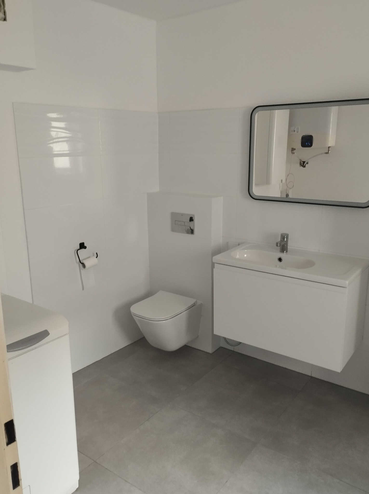 Nowoczesna, minimalistyczna łazienka w odcieniach bieli i szarości, z podwieszaną toaletą, szafką pod umywalką i lustrem w czarnej ramie, widoczna pralka z boku kadru.