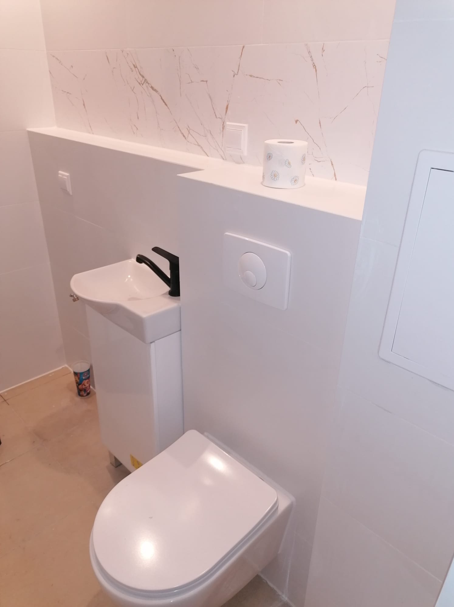 Nowoczesna toaleta z umywalką nablatową i czarną armaturą, białe płytki imitujące marmur na ścianach, widoczny pojemnik na szczoteczki dla dzieci.