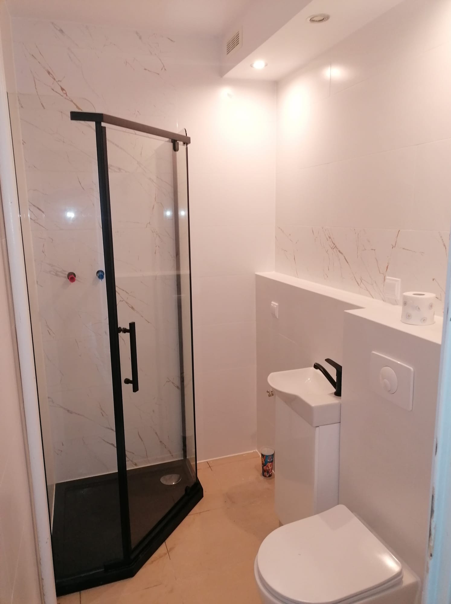 Nowoczesna łazienka z kabiną prysznicową z czarną ramą, białą umywalką na postumencie i toaletą, wykończona płytkami imitującymi marmur.