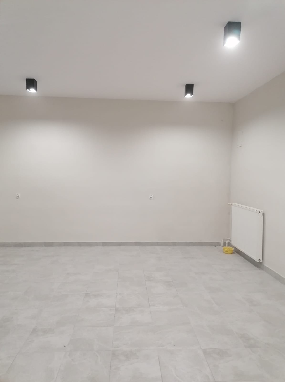 Pusty, świeżo wykończony pokój z szarymi płytkami podłogowymi, trzema czarnymi lampami sufitowymi, białymi ścianami i grzejnikiem na ścianie. Żółta miska stoi pod grzejnikiem.