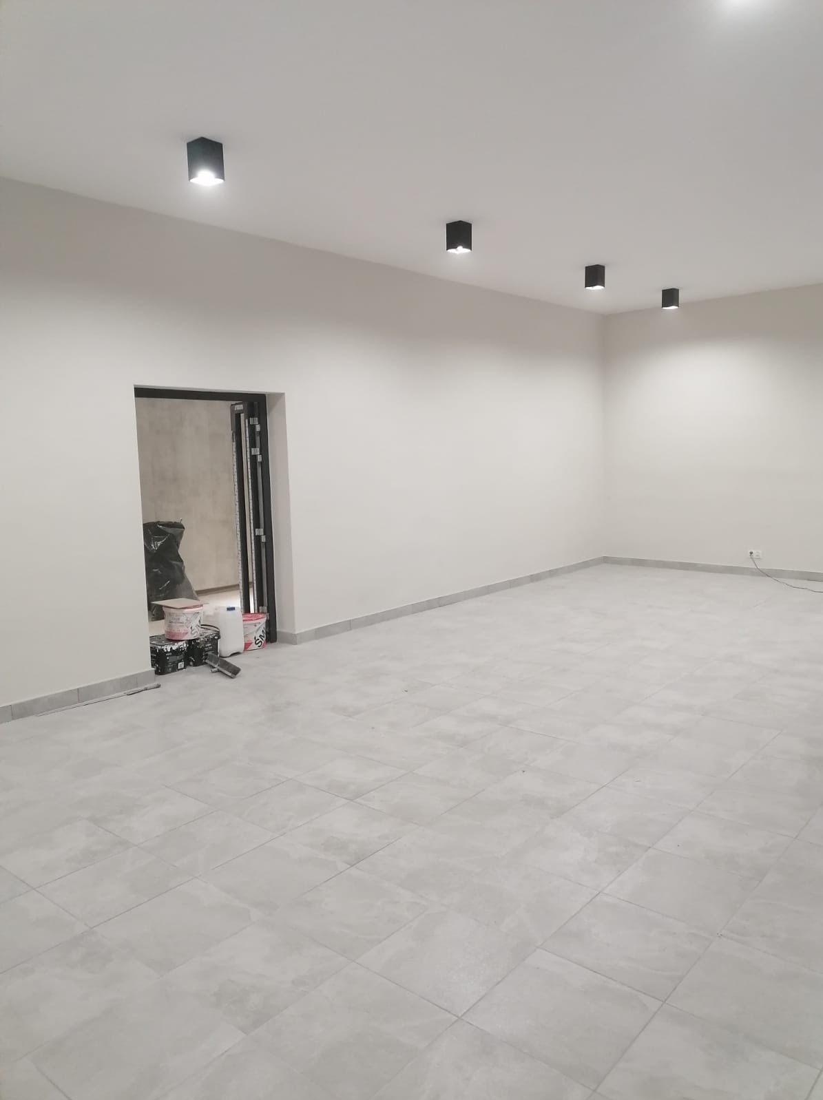 Wykończone wnętrze: szare płytki podłogowe, świeżo pomalowane białe ściany z czarnymi reflektorami sufitowymi, widoczne puszki z farbą i narzędzia malarskie przy wejściu.