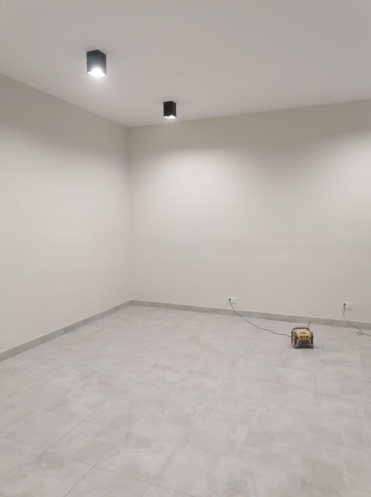 Puste pomieszczenie po remoncie z szarymi płytkami podłogowymi i listwami przypodłogowymi, dwa czarne reflektory sufitowe, żółty radioodbiornik DeWalt na podłodze podłączony do gniazdka elektrycznego.