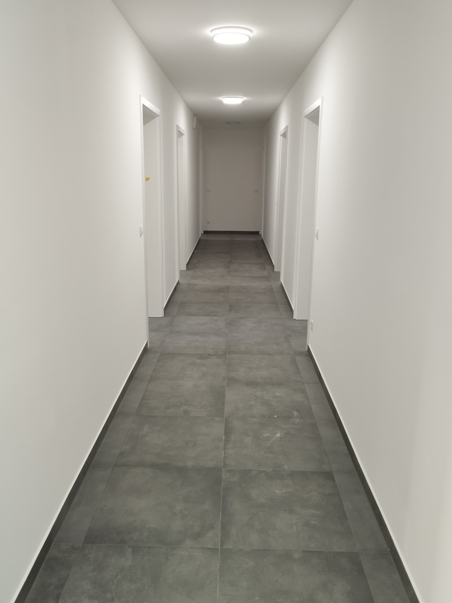 Długi, jasny korytarz z szarymi, kwadratowymi płytkami podłogowymi i białymi ścianami z białymi drzwiami po obu stronach, oświetlony trzema okrągłymi lampami sufitowymi.