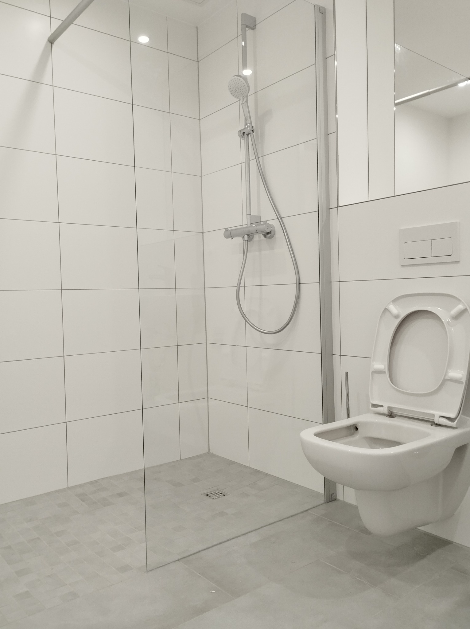 Minimalistyczna, jasna łazienka z kabiną prysznicową typu walk-in, wiszącą toaletą z otwartą klapą i białymi płytkami na ścianach.