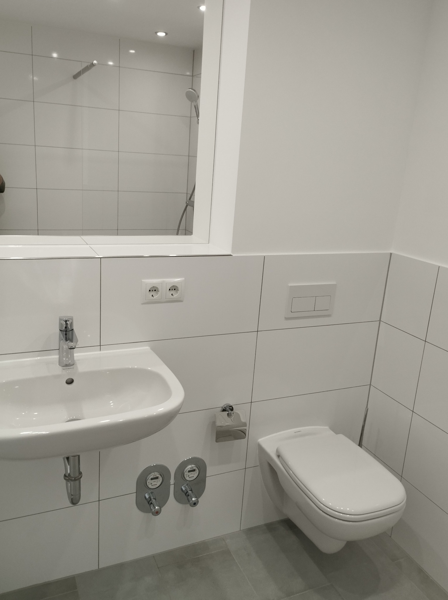 Nowoczesna, minimalistyczna łazienka w bieli z wiszącą toaletą, umywalką i dużym lustrem, widoczne szare płytki podłogowe i chromowana armatura.