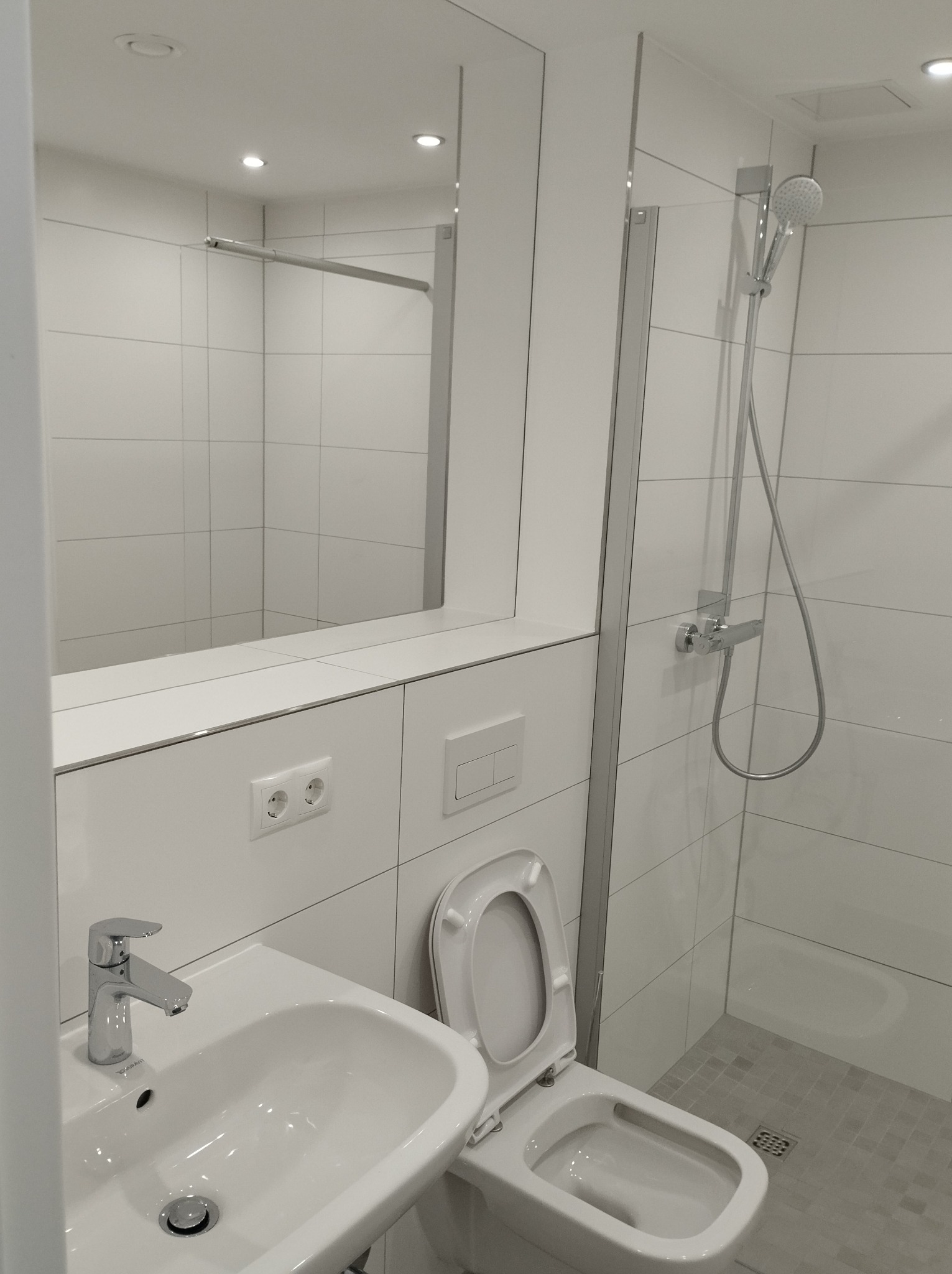 Nowoczesna, jasna łazienka z białymi płytkami, umywalką z chromowaną baterią, toaletą z otwartą klapą i kabiną prysznicową ze szklanymi drzwiami.