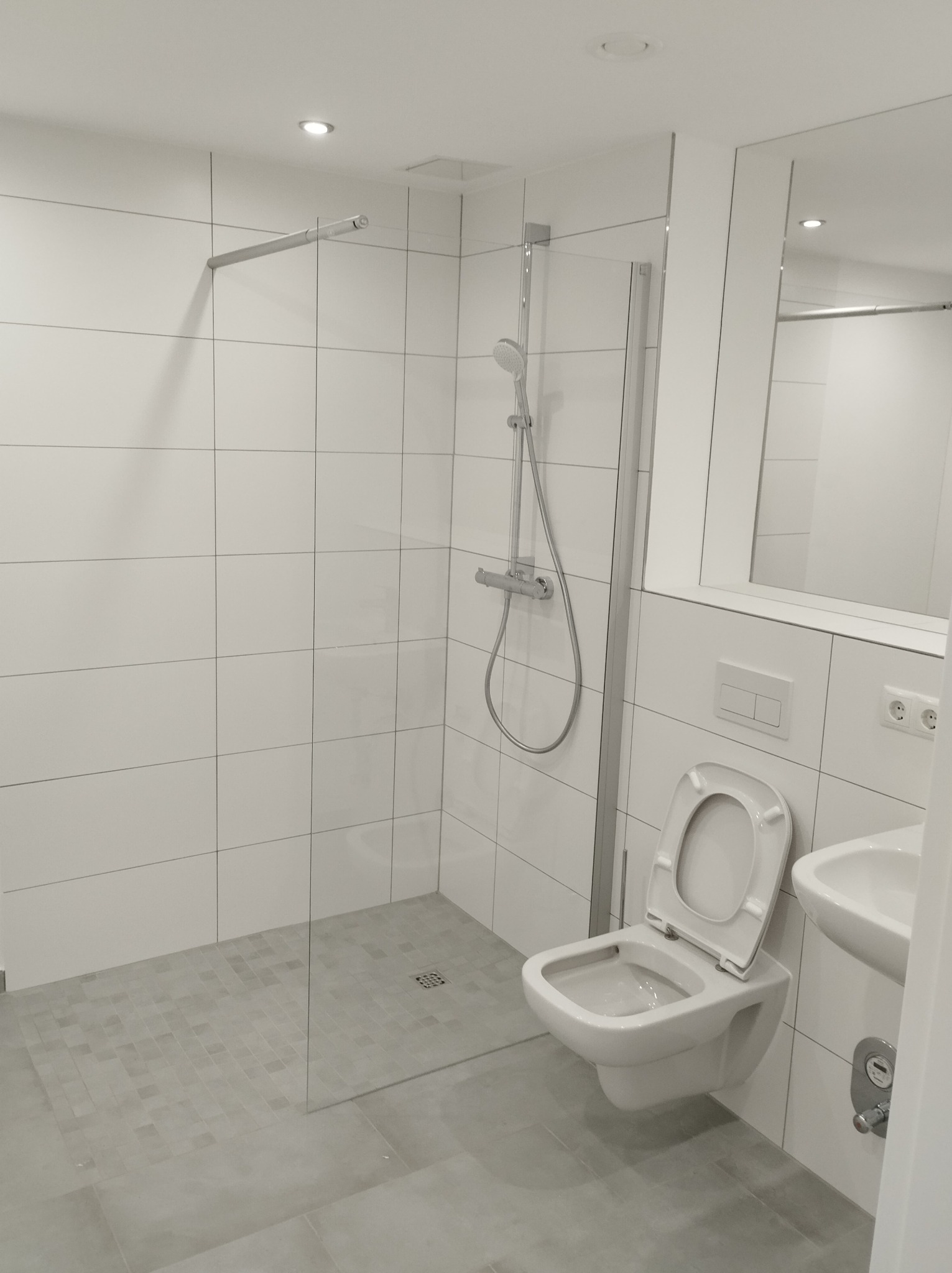 Nowoczesna łazienka z kabiną prysznicową typu walk-in, wykończona białymi płytkami ściennymi i szarymi płytkami podłogowymi w strefie prysznica, z widoczną toaletą wiszącą i umywalką.