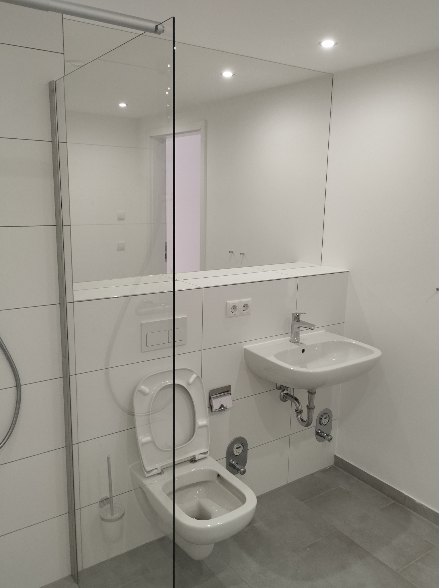 Nowoczesna, minimalistyczna łazienka z białymi płytkami, wiszącą toaletą z otwartą klapą, umywalką i szklaną kabiną prysznicową, oświetlona punktowymi lampami sufitowymi.