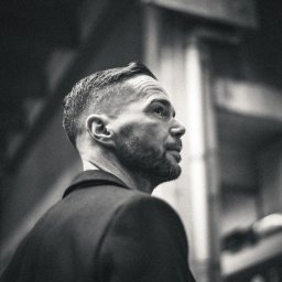 Adrian Krać Fotografia - Czarno-białe zdjęcie profilowe mężczyzny z brodą, w garniturze, patrzącego w górę, z wyraźnym oświetleniem i fakturą skóry.
