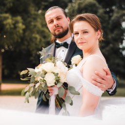 Adrian Krać Fotografia - Para młoda w eleganckich strojach pozuje z bukietem białych róż i zielonych liści na tle zieleni parkowej, z delikatnym uśmiechem skierowanym w stronę obiektywu.