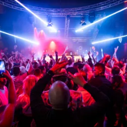 Tłum ludzi tańczących w klubie nocnym, oświetlonym neonami i światłami scenicznymi, z podniesionymi rękami i telefonami w górze.