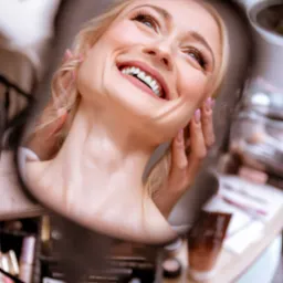 Uśmiechnięta blond kobieta spogląda w górę, odbicie w lustrze otoczonym akcesoriami do makijażu na toaletce.