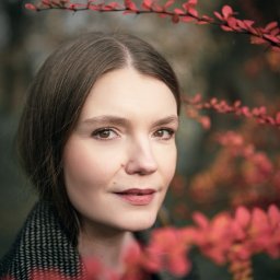 Adrian Krać Fotografia - Portret kobiety o jasnej karnacji, otoczonej gałązkami z czerwonymi liśćmi, w jesiennej scenerii. Ubrana w płaszcz w pepitkę, z delikatnym uśmiechem.