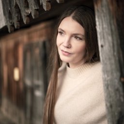 Adrian Krać Fotografia - Portret kobiety o długich, brązowych włosach, w beżowym swetrze, spoglądającej w prawo, w drewnianej ramie z elementami rustykalnymi w tle.