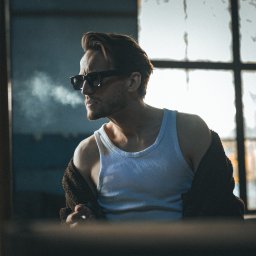 Adrian Krać Fotografia - Mężczyzna w okularach przeciwsłonecznych, w białym podkoszulku, wypuszcza dym na tle okna z widocznymi jesiennymi liśćmi. Ujęcie w stylu vintage, z ciemną tonacją.