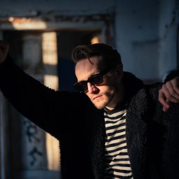 Adrian Krać Fotografia - Mężczyzna w okularach przeciwsłonecznych, oparty o framugę drzwi w opuszczonym budynku, oświetlony mocnym światłem słonecznym, w czarno-białej koszulce i czarnym płaszczu.