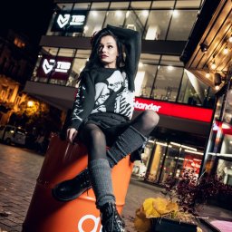 Adrian Krać Fotografia - Stylowa sesja: młoda kobieta w czarnym swetrze z motywem czaszek pozuje siedząc na pomarańczowej beczce na tle nocnego miasta z oświetlonym budynkiem galerii.