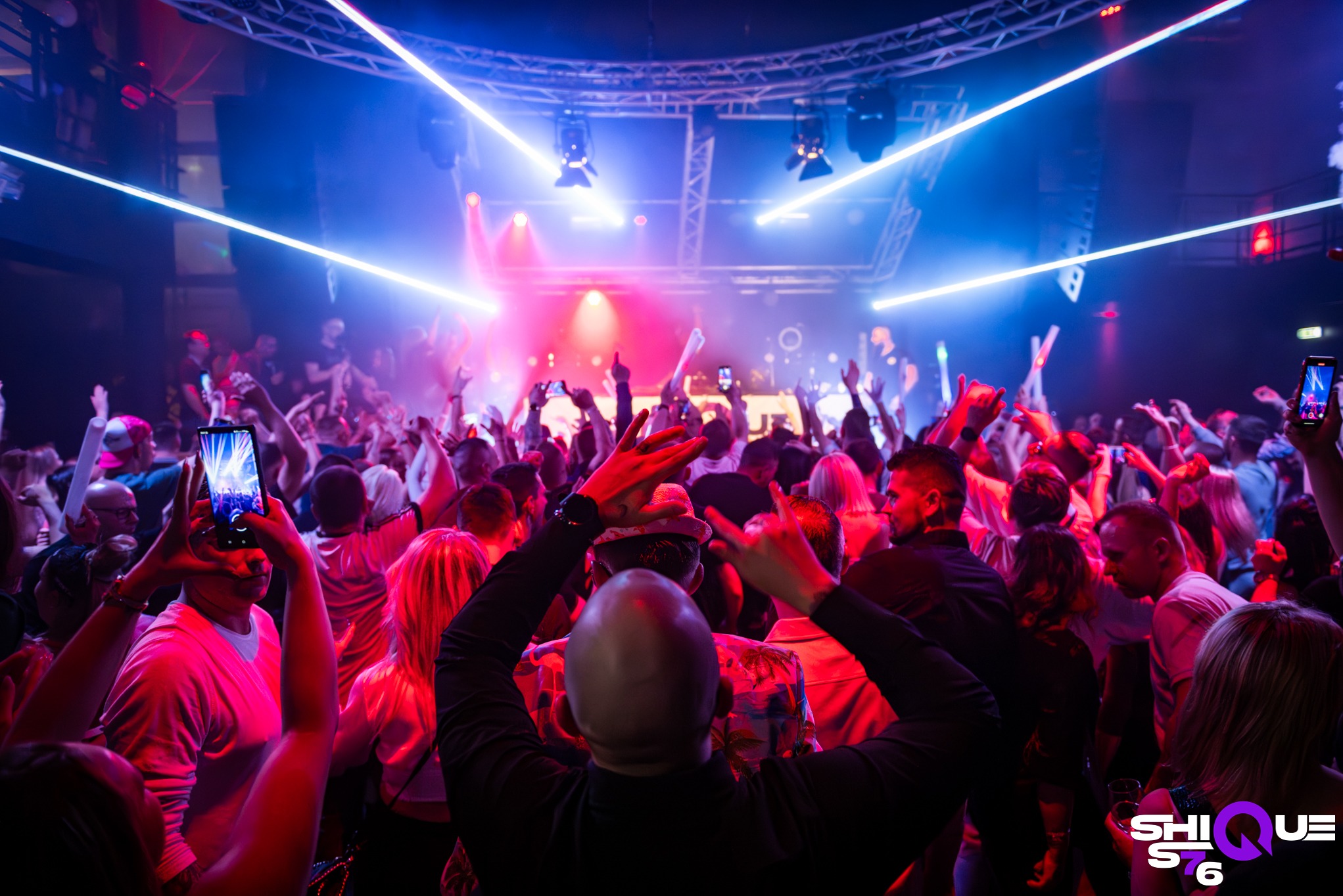 Tłum ludzi tańczących w klubie nocnym, oświetlonym neonami i światłami scenicznymi, z podniesionymi rękami i telefonami w górze.