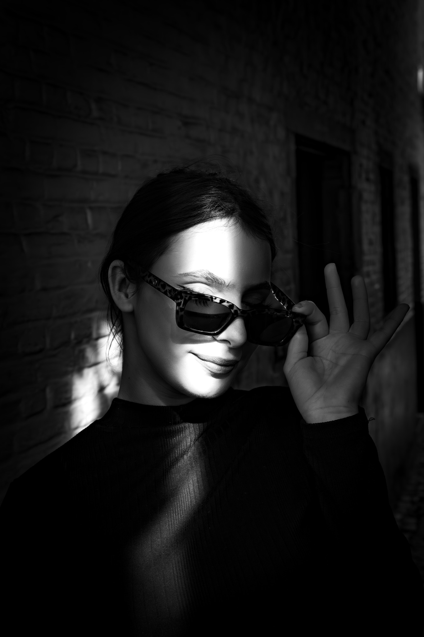 Portret kobiety w czerni, częściowo oświetlonej promieniami słońca, z okularami w panterkę, na tle ceglanej ściany.