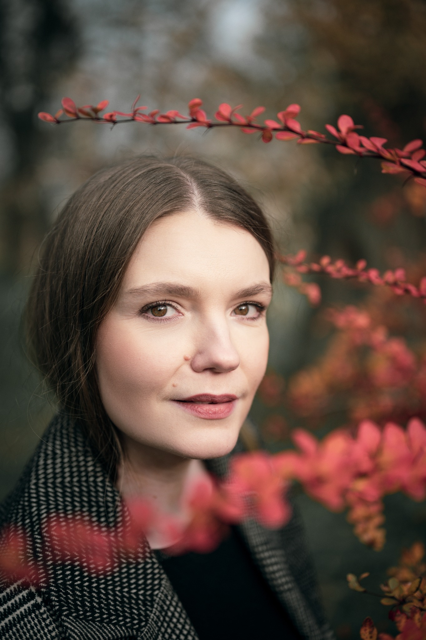 Portret kobiety o jasnej karnacji, otoczonej gałązkami z czerwonymi liśćmi, w jesiennej scenerii. Ubrana w płaszcz w pepitkę, z delikatnym uśmiechem.