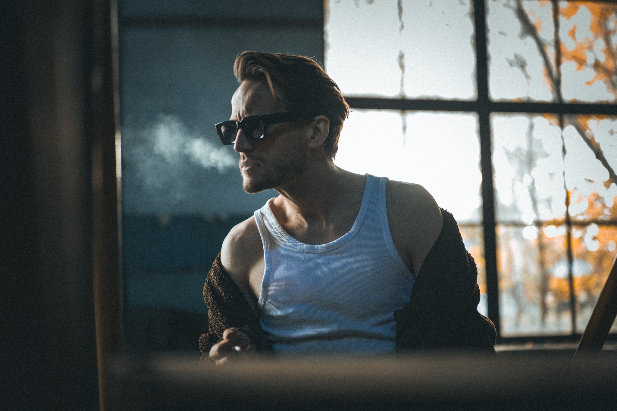 Mężczyzna w okularach przeciwsłonecznych, w białym podkoszulku, wypuszcza dym na tle okna z widocznymi jesiennymi liśćmi. Ujęcie w stylu vintage, z ciemną tonacją.