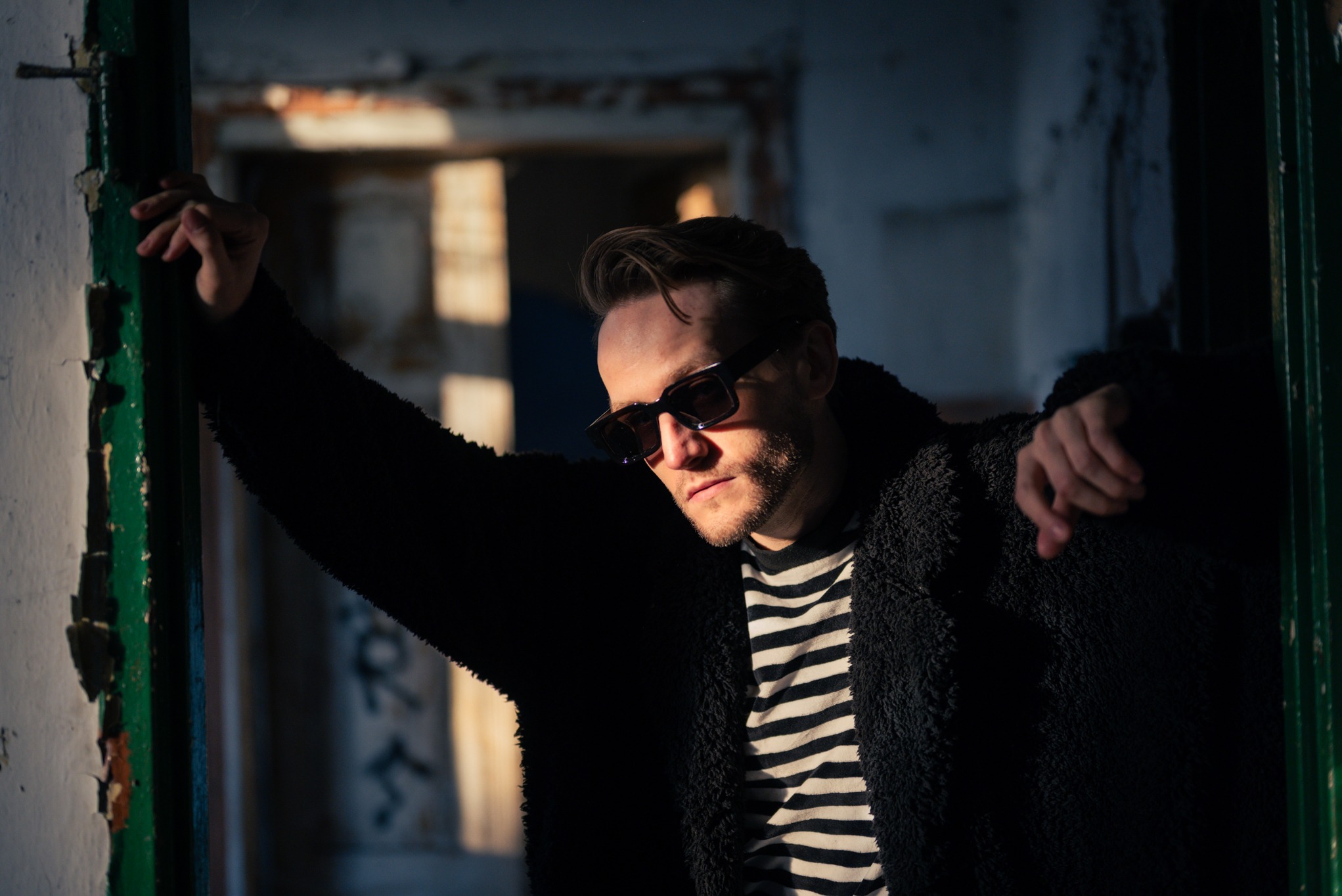 Mężczyzna w okularach przeciwsłonecznych, oparty o framugę drzwi w opuszczonym budynku, oświetlony mocnym światłem słonecznym, w czarno-białej koszulce i czarnym płaszczu.