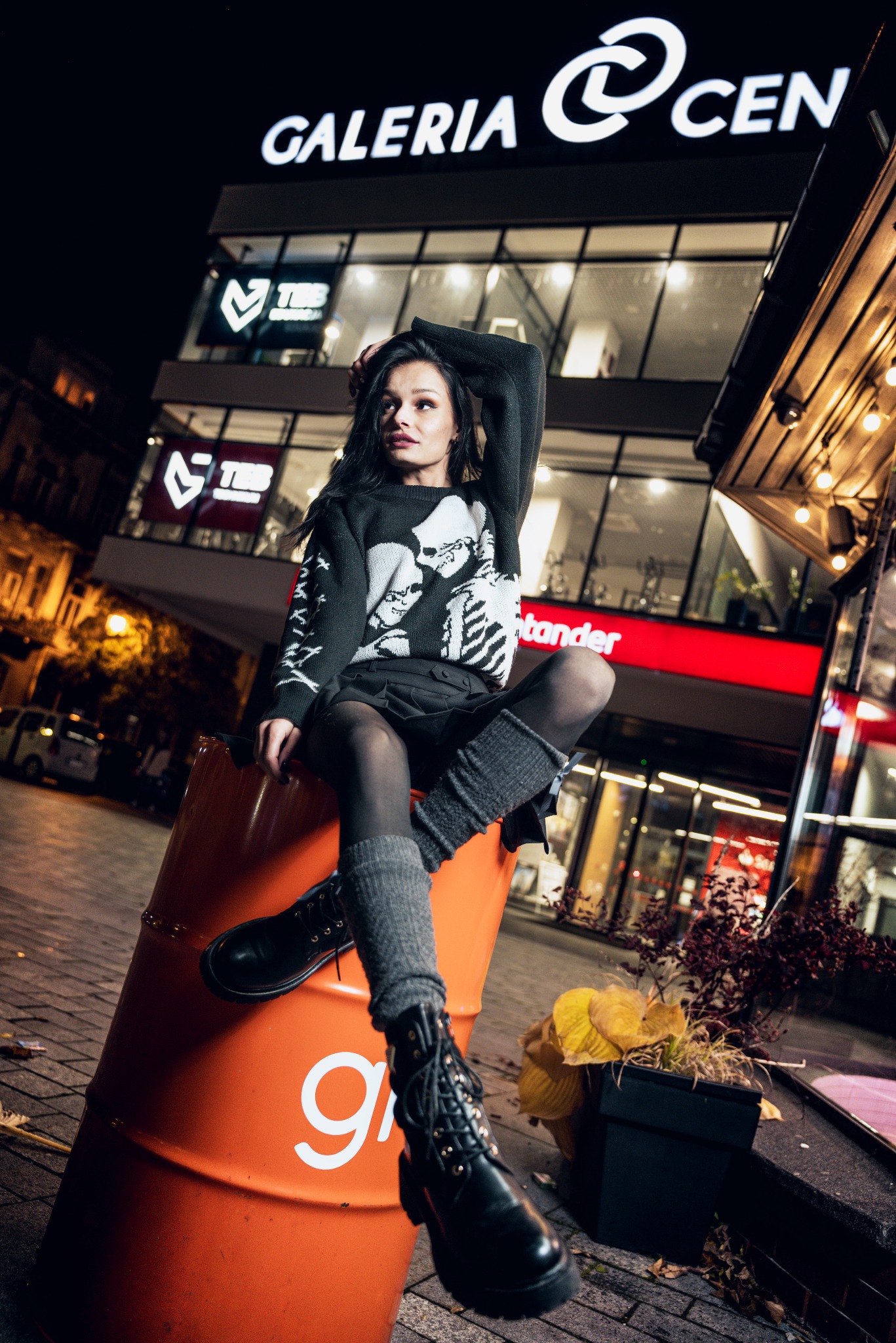 Stylowa sesja: młoda kobieta w czarnym swetrze z motywem czaszek pozuje siedząc na pomarańczowej beczce na tle nocnego miasta z oświetlonym budynkiem galerii.