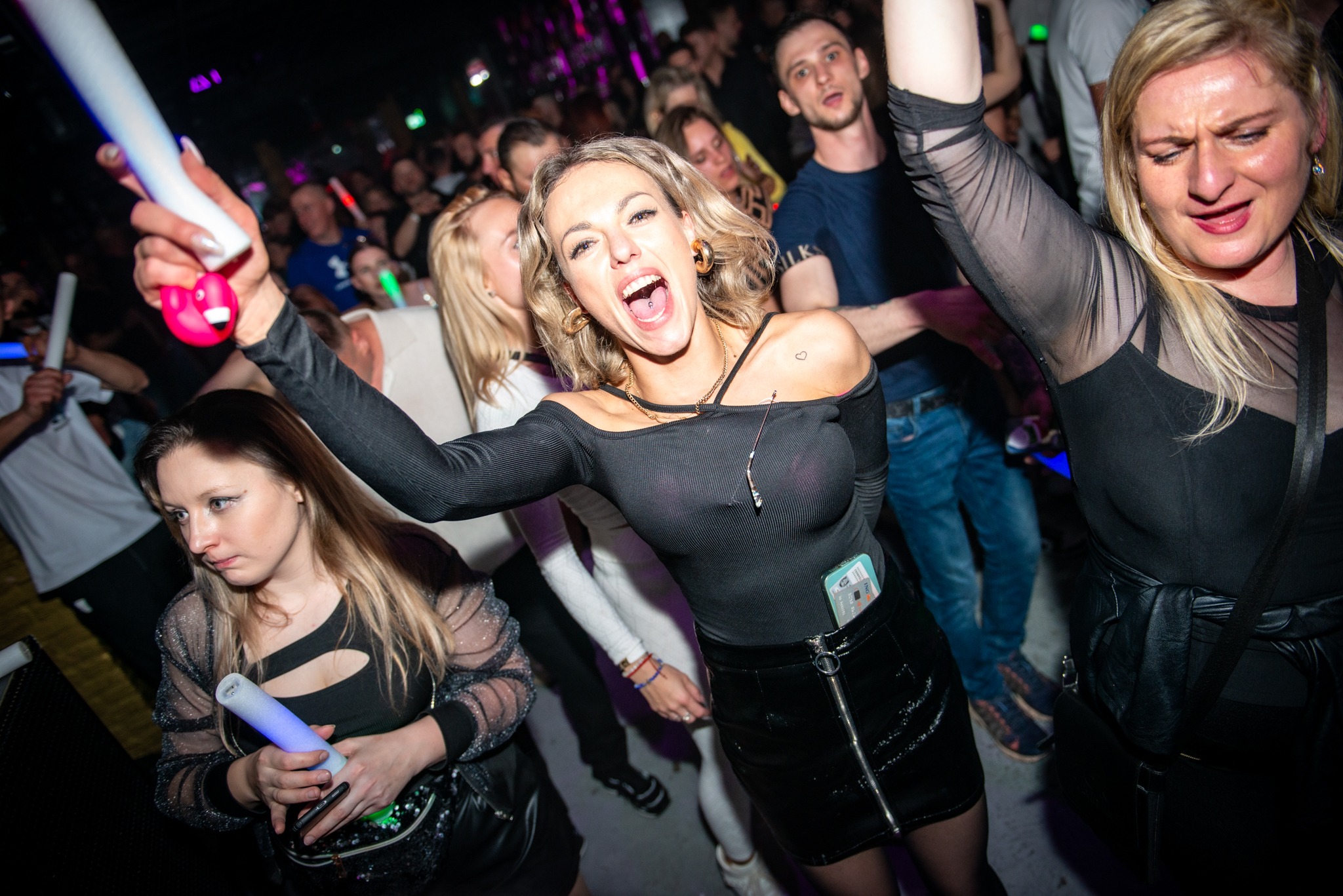 Rozbawiona blondynka z otwartymi ustami na parkiecie, w tle tłum ludzi z neonowymi pałeczkami, dynamiczne ujęcie z imprezy w klubie nocnym.