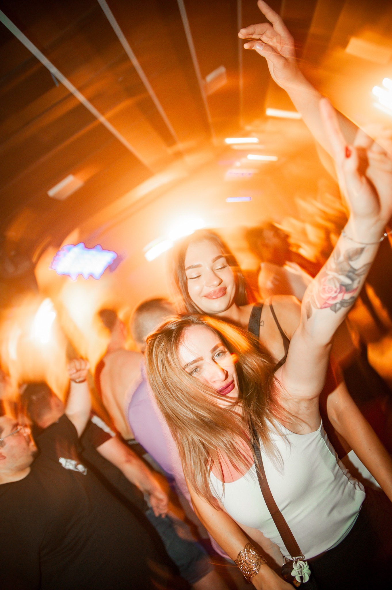 Rozmazane ujęcie dwóch uśmiechniętych kobiet na parkiecie, w tle rozświetlona scena i bawiący się ludzie. Widoczne tatuaże i biżuteria. Dynamiczne, pomarańczowe oświetlenie.