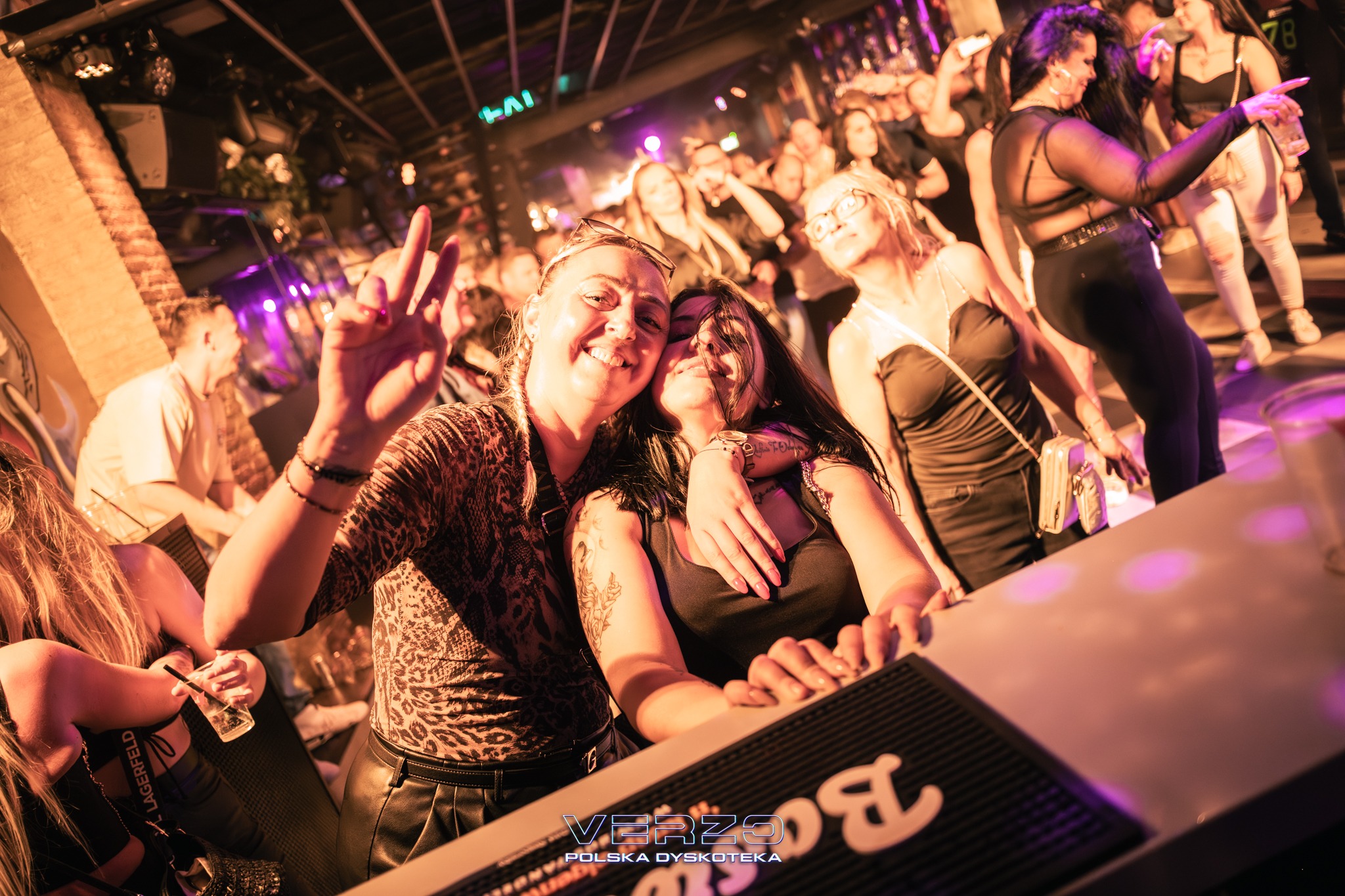 Uśmiechnięte kobiety pozują w dyskotece Verzo, oświetlone fioletowym światłem. W tle tańczący tłum i ceglany filar. Jedna z kobiet pokazuje znak zwycięstwa.