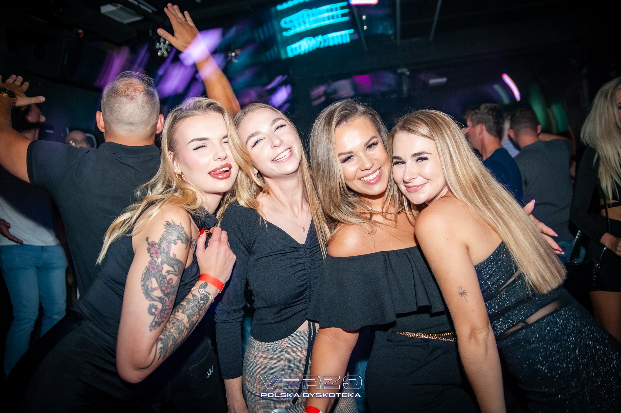 Uśmiechnięte kobiety w klubie nocnym Verzo, Polska Dyskoteka, z neonowym oświetleniem w tle. Jedna z kobiet ma tatuaż smoka na ramieniu.