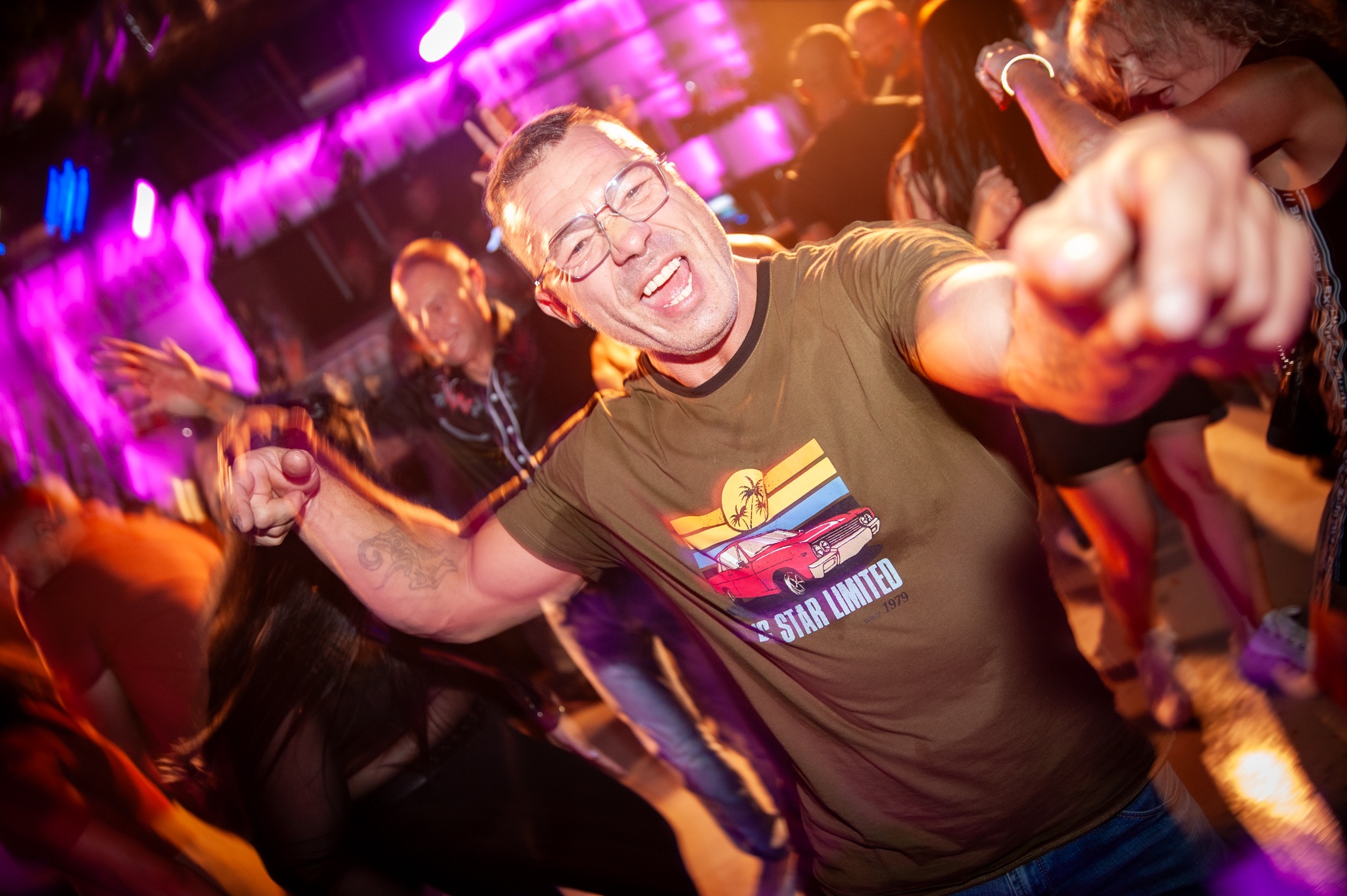 Rozbawiony mężczyzna w okularach i koszulce z nadrukiem tańczy na parkiecie w klubie, otoczony przez rozmazane sylwetki innych bawiących się osób, oświetlony fioletowymi światłami.