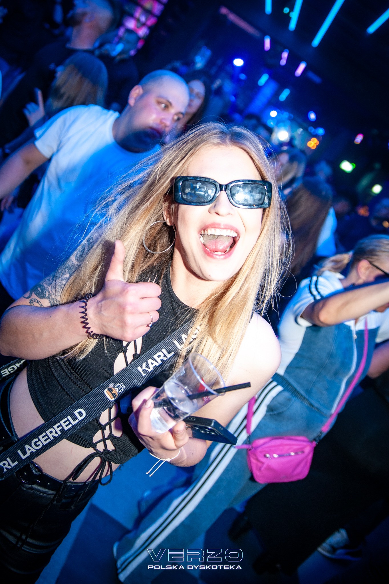 Uśmiechnięta blondynka w okularach na imprezie, z drinkiem i kciukiem w górę, w tle rozmazane sylwetki i światła dyskoteki. Dynamiczne ujęcie z dołu.