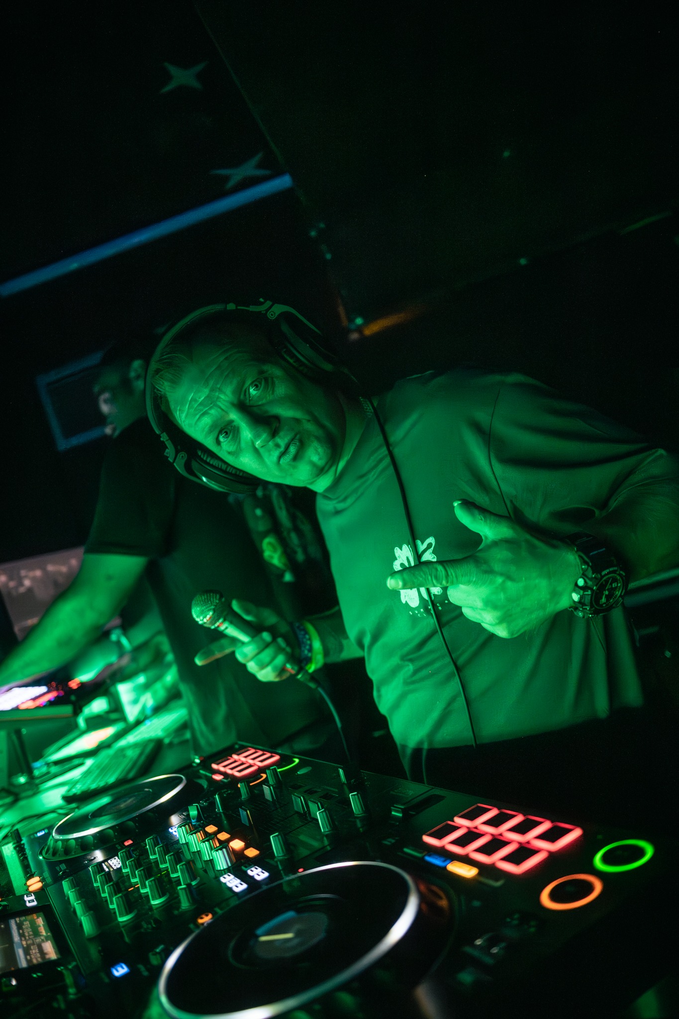 DJ w zielonym oświetleniu, z mikrofonem i słuchawkami, prezentuje sprzęt DJ-ski. Drugi DJ w tle. Scena klubowa, dynamiczne ujęcie z bliska, ciemne tło z elementami dekoracji.