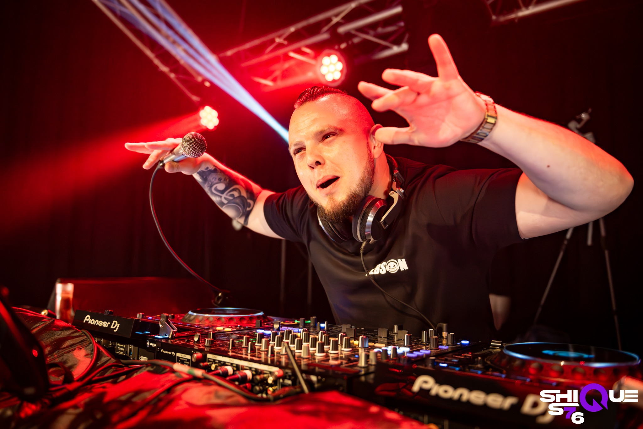 DJ z tatuażem, w czarnej koszulce, z mikrofonem i słuchawkami, grający na sprzęcie Pioneer DJ w czerwonym oświetleniu scenicznym podczas występu.