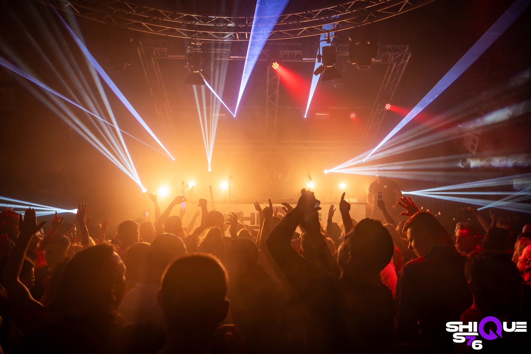 Rozświetlony tłum na imprezie z podniesionymi rękami, oświetlony przez reflektory i lasery. Widoczna scena i konstrukcja oświetleniowa.