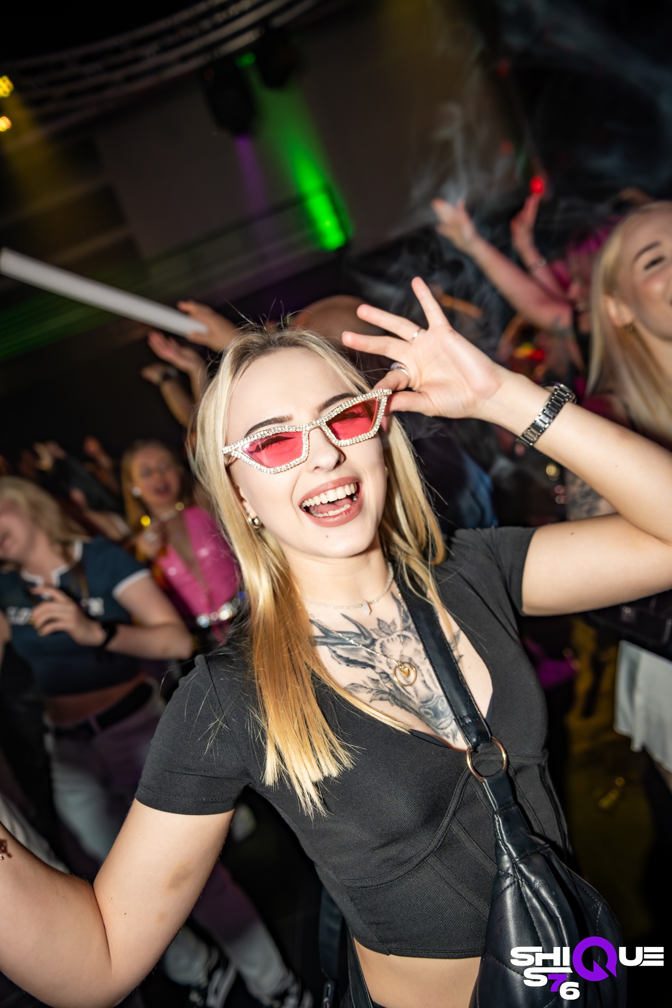 Uśmiechnięta blondynka w różowych okularach na imprezie, z tatuażem na dekolcie, w tle rozbawiony tłum i efekty świetlne. Dynamiczne ujęcie z bliska.