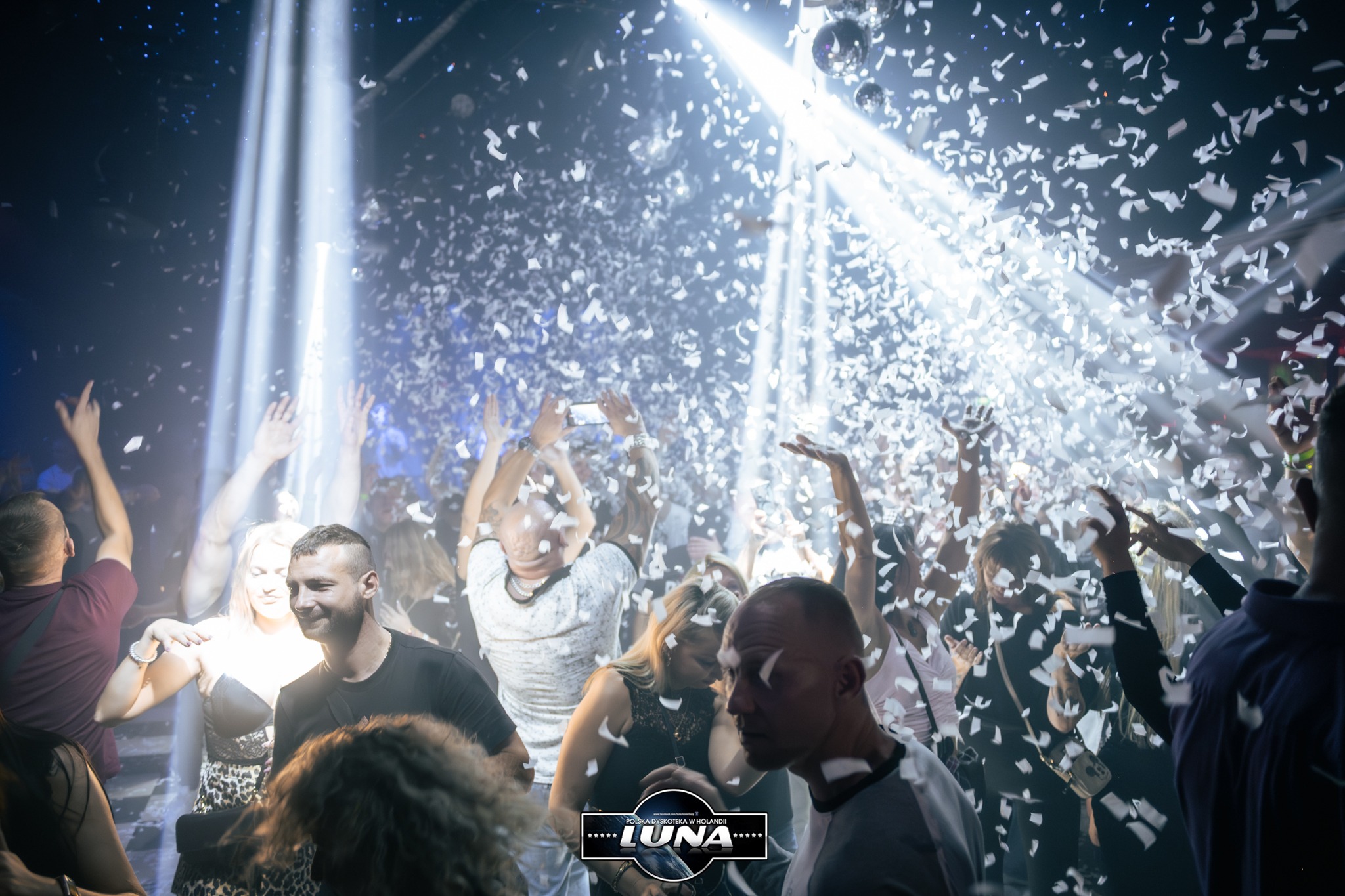 Rozświetlona sala dyskoteki z bawiącymi się ludźmi, konfetti w powietrzu i logo klubu Luna w dolnej części kadru. Dynamiczne ujęcie z imprezy.